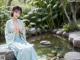 A woman in a blue dress sitting on a rock by a pond.