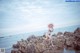 A woman in a bikini and straw hat standing on rocks by the ocean.